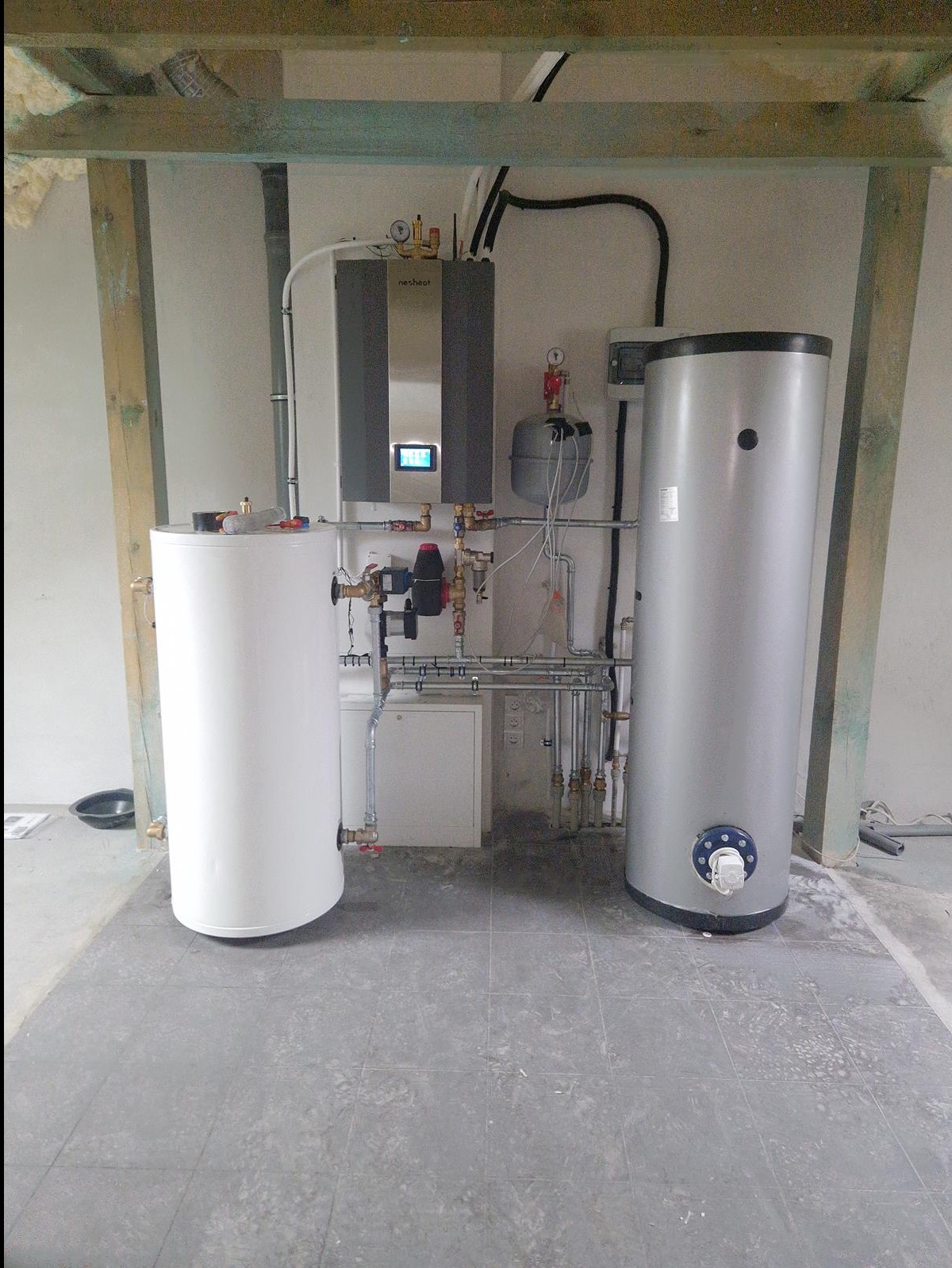 Dwa zbiorniki na wodę (biały i szary) oraz jednostka grzewcza zamontowane w surowym pomieszczeniu z widocznymi instalacjami hydraulicznymi i elektrycznymi, wskazujące na etap montażu.