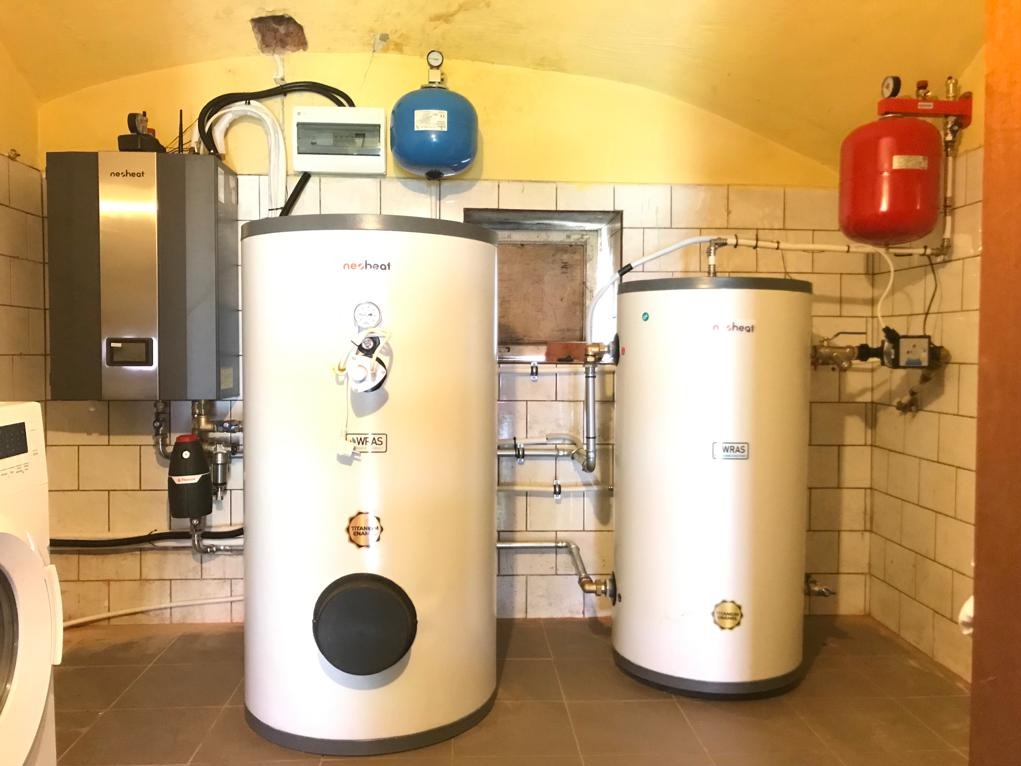 Instalacja dwóch białych zbiorników buforowych Nesheat z osprzętem, widoczny także szary piec, instalacja elektryczna i pralka w pomieszczeniu wyłożonym kafelkami.