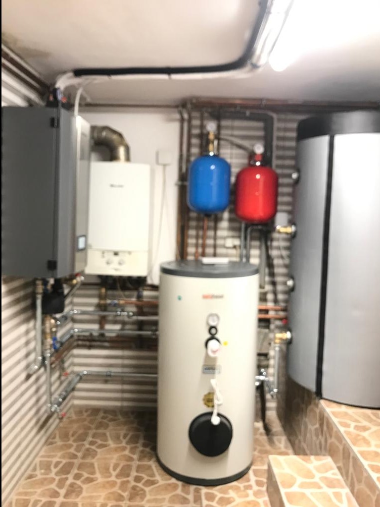 Kotłownia z zainstalowanym zasobnikiem ciepłej wody użytkowej, widoczne rury miedziane, zbiorniki wyrównawcze w kolorze niebieskim i czerwonym, oraz płytki ceramiczne na podłodze.