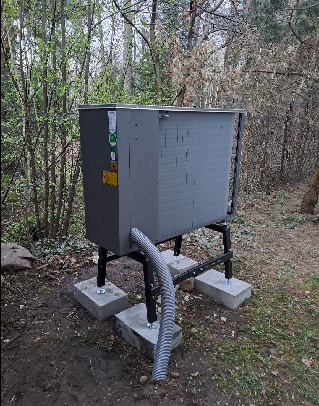 Szara, zewnętrzna jednostka urządzenia na betonowych podstawach w ogrodzie, z widocznym karbowanym wężem odprowadzającym