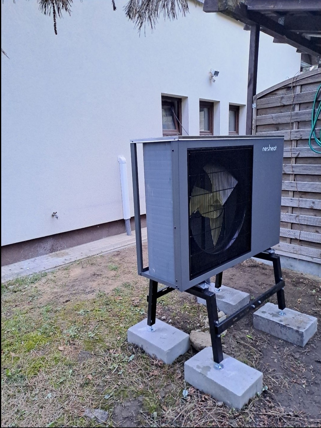 Szara jednostka zewnętrzna pompy ciepła marki Nesheat, zamontowana na czarnej, metalowej konstrukcji wspartej na betonowych bloczkach, na tle elewacji budynku i drewnianego płotu.