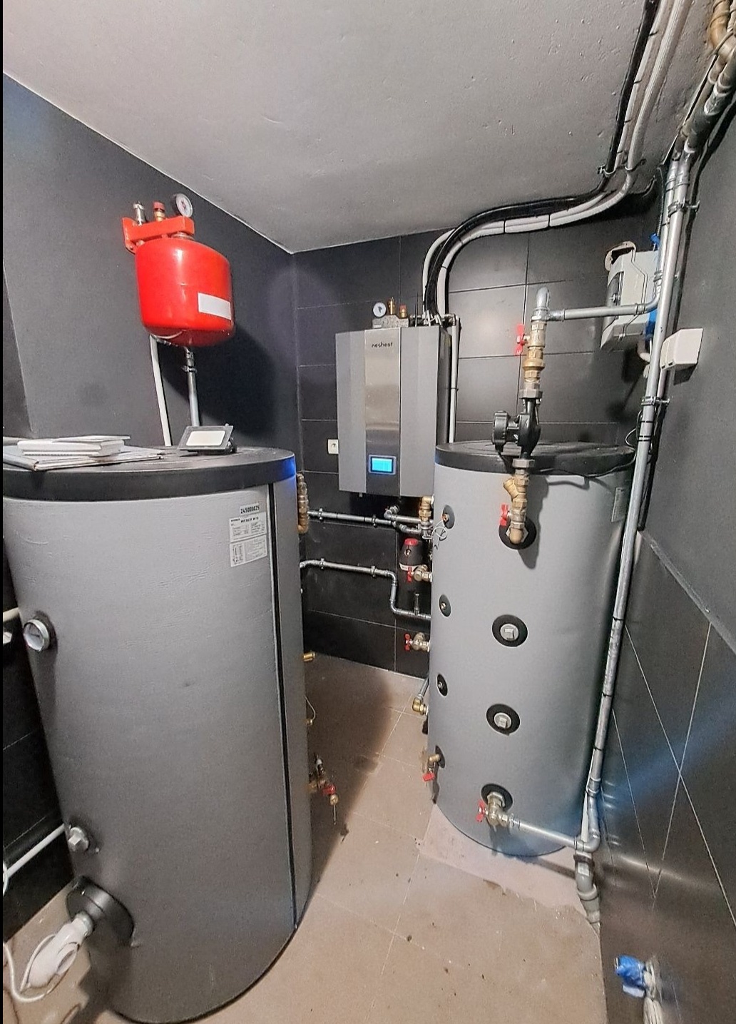 Zestaw urządzeń w kotłowni: dwa szare zbiorniki akumulacyjne, srebrny piec z niebieskim wyświetlaczem marki Nesheat, czerwony zbiornik wyrównawczy, instalacja rurowa z zaworami, czarne płytki...