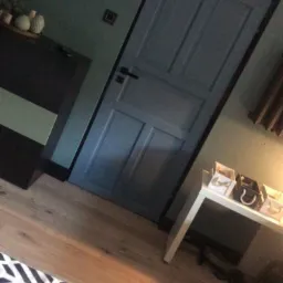 Pomalowane na jednolity, ciemny odcień zieleni ściany i niebieskie drzwi w pokoju z parkietem i minimalistycznym umeblowaniem.