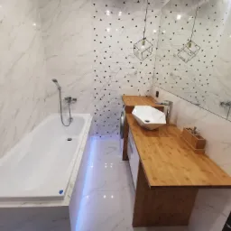 Nowoczesna łazienka z biało-marmurowymi płytkami, drewnianym blatem pod umywalkę i geometrycznym wzorem na ścianie, oświetlona wiszącymi lampami w kształcie sześcianów.