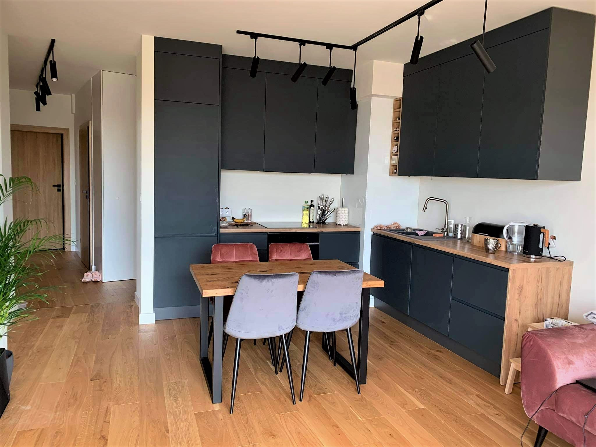 Nowoczesna kuchnia z ciemnymi szafkami, drewnianym blatem i stołem z szarymi krzesłami, oświetlona reflektorami sufitowymi