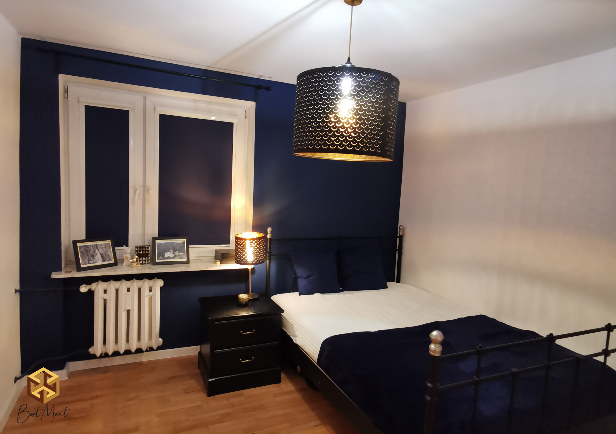 Sypialnia z ciemnoniebieskimi ścianami, białym oknem i grzejnikiem, czarną metalową ramą łóżka i lampą z ażurowym kloszem w kolorze czarno-złotym.
