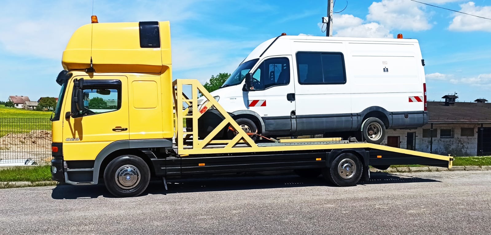 Żółta ciężarówka z platformą przewozi białego busa na tle błękitnego nieba i zieleni.