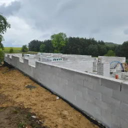 Wznoszenie ścian fundamentowych z bloczków betonowych H+H. Widoczne palety z bloczkami i częściowo zrealizowana konstrukcja na tle zieleni i pochmurnego nieba.