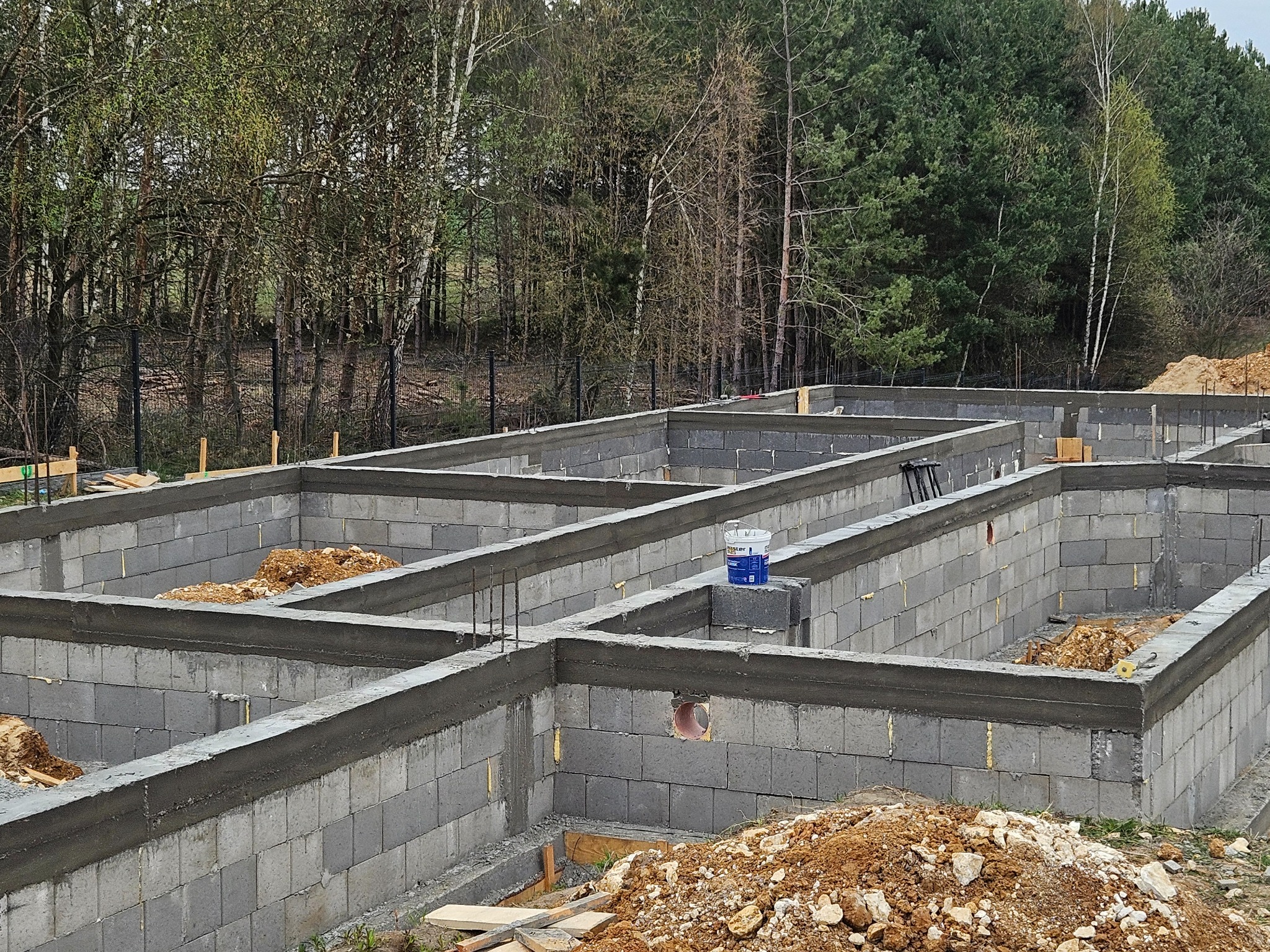 Fundamenty domu z bloczków betonowych, zalane betonem, z widoczną puszką farby, na tle zielonego lasu i drzew. Widok z góry, oświetlenie dzienne.