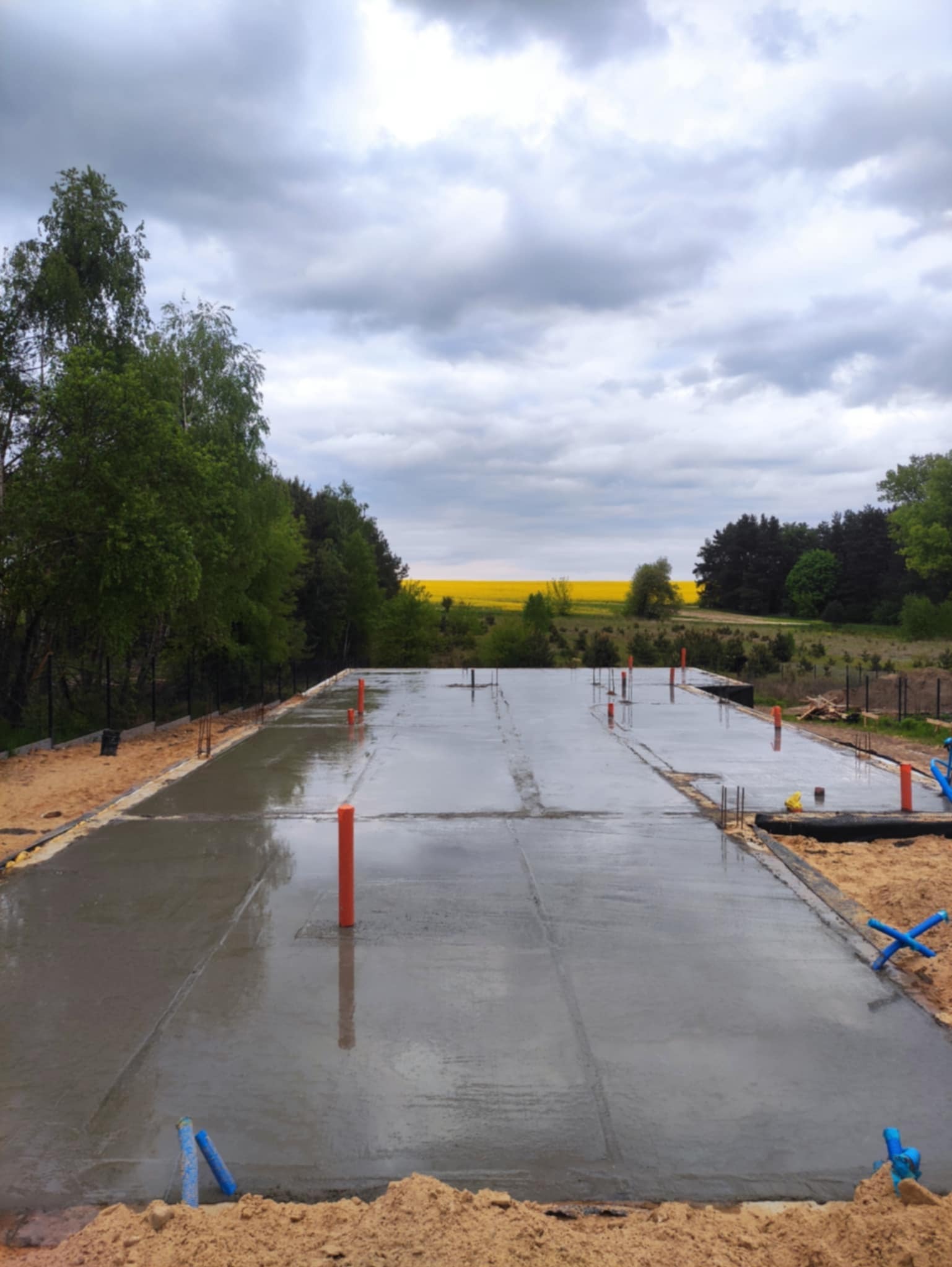 Świeżo wylana płyta fundamentowa z wystającymi rurami instalacyjnymi, otoczona piaskiem, na tle zieleni i żółtego pola pod pochmurnym niebem.