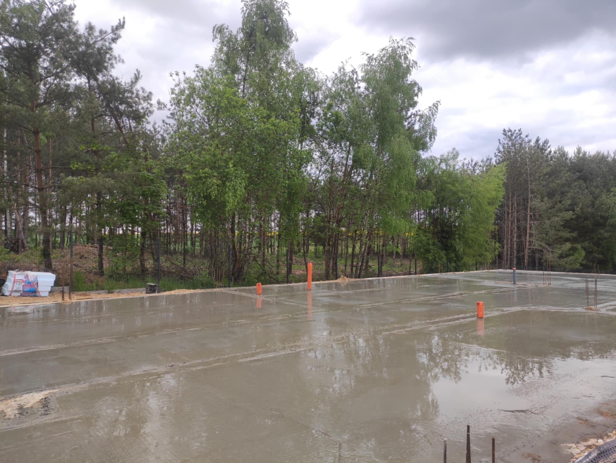 Świeżo wylana, gładka betonowa płyta fundamentowa z widocznymi wystającymi prętami zbrojeniowymi i pomarańczowymi rurami instalacyjnymi, otoczona zielenią drzew.