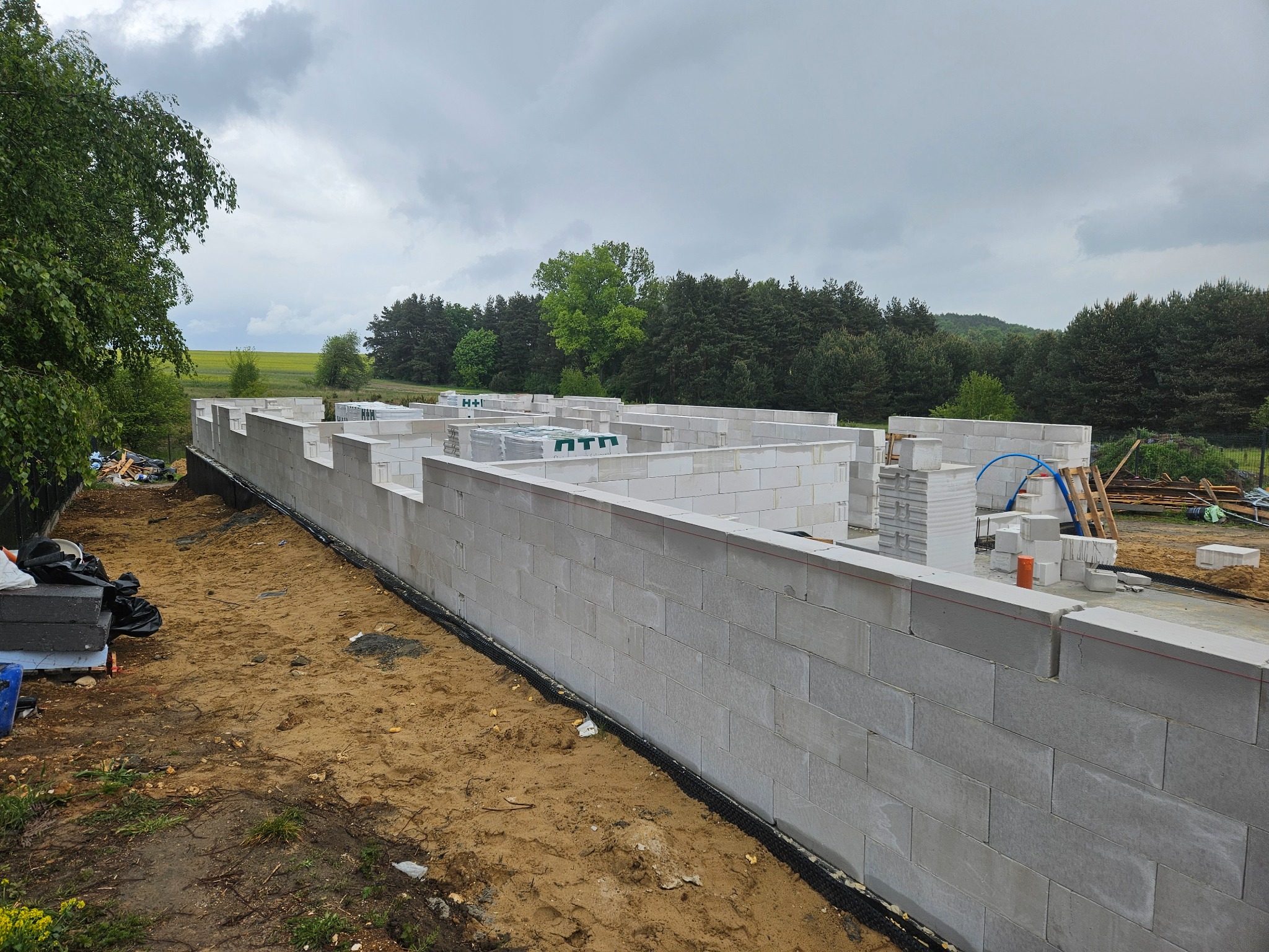 Wznoszenie ścian fundamentowych z bloczków betonowych H+H. Widoczne palety z bloczkami i częściowo zrealizowana konstrukcja na tle zieleni i pochmurnego nieba.
