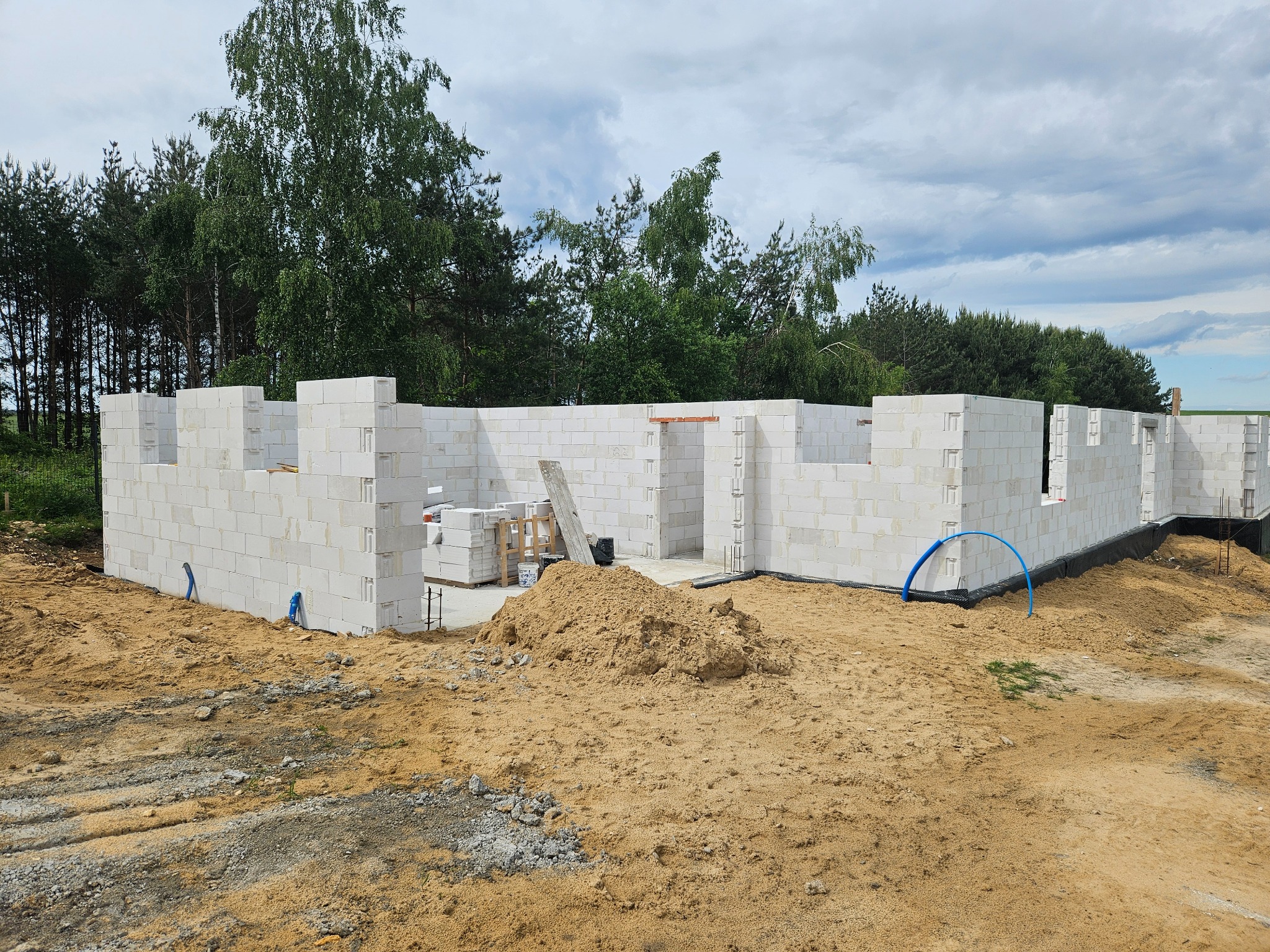 Budowa domu w toku: ściany z białych bloczków, widoczne niebieskie rury, piasek i zieleń w tle. Świeża konstrukcja w otoczeniu natury.