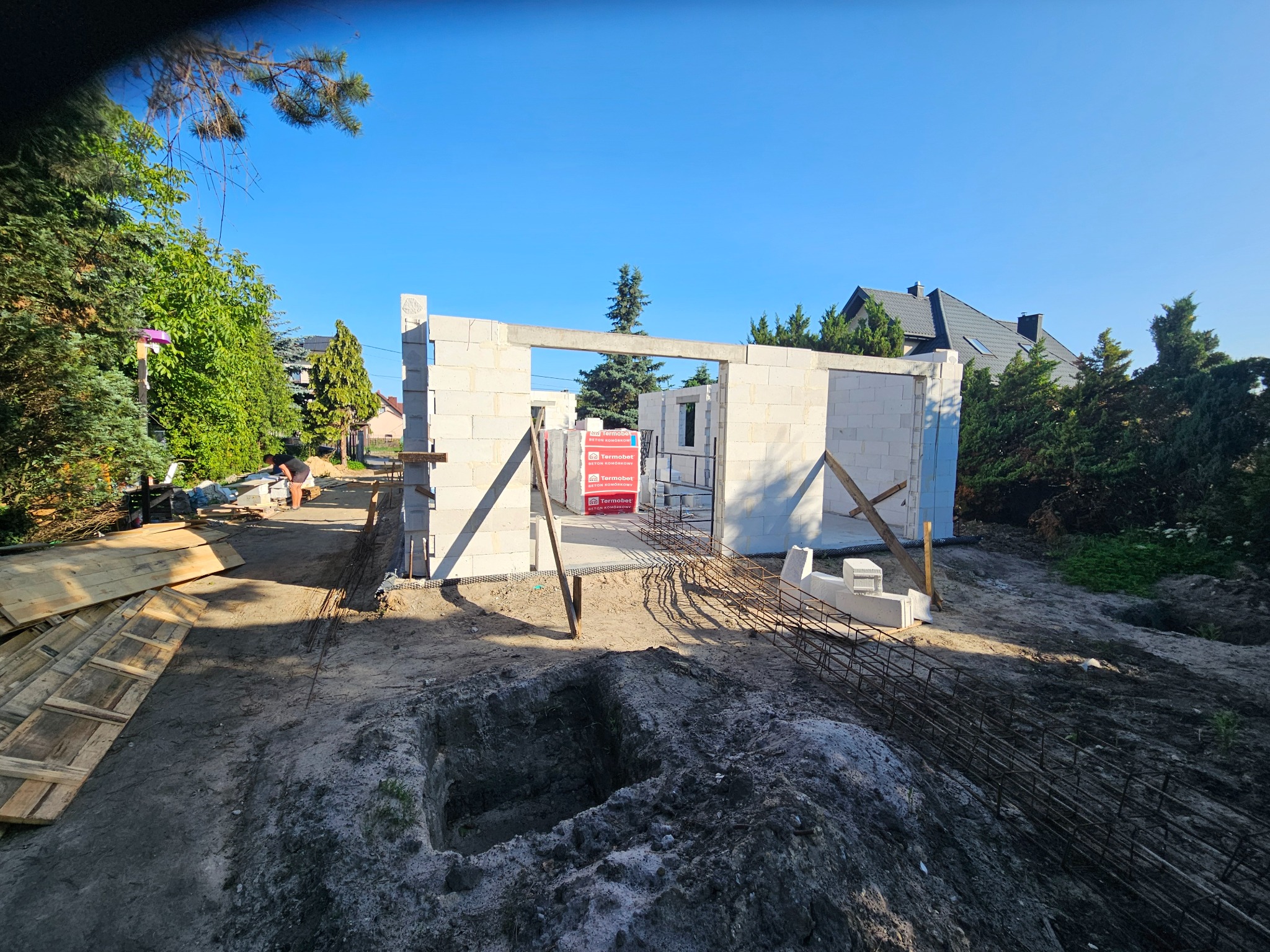 Budowa domu: ściany z betonu komórkowego wzniesione, widoczne zbrojenia i palety z bloczkami Termobet. W tle zieleń i błękitne niebo.