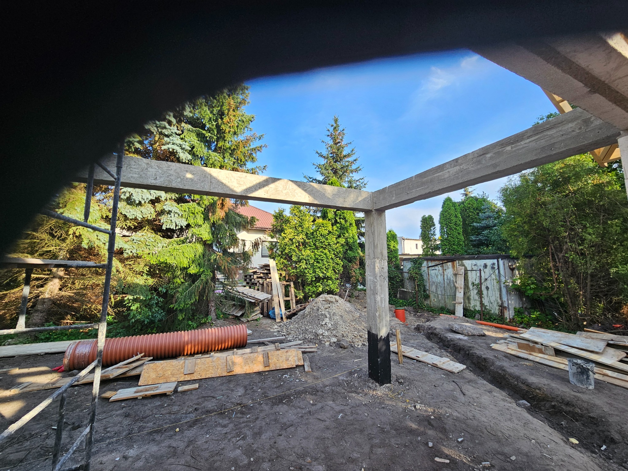 Surowa konstrukcja domu w trakcie budowy: betonowe słupy i belki na tle zieleni, wykopane fundamenty, rura i drewniane deski na placu budowy w słoneczny dzień.