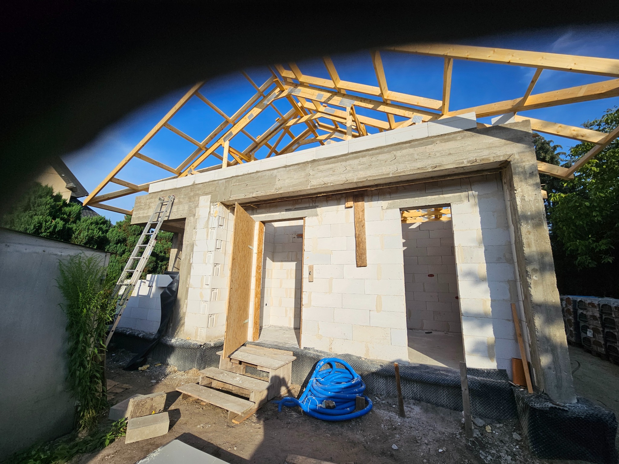 Budowa domu: ściany z białych bloczków, drewniana konstrukcja dachu w trakcie montażu, niebieski wąż na ziemi, drabina oparta o ścianę. Widok z zewnątrz.