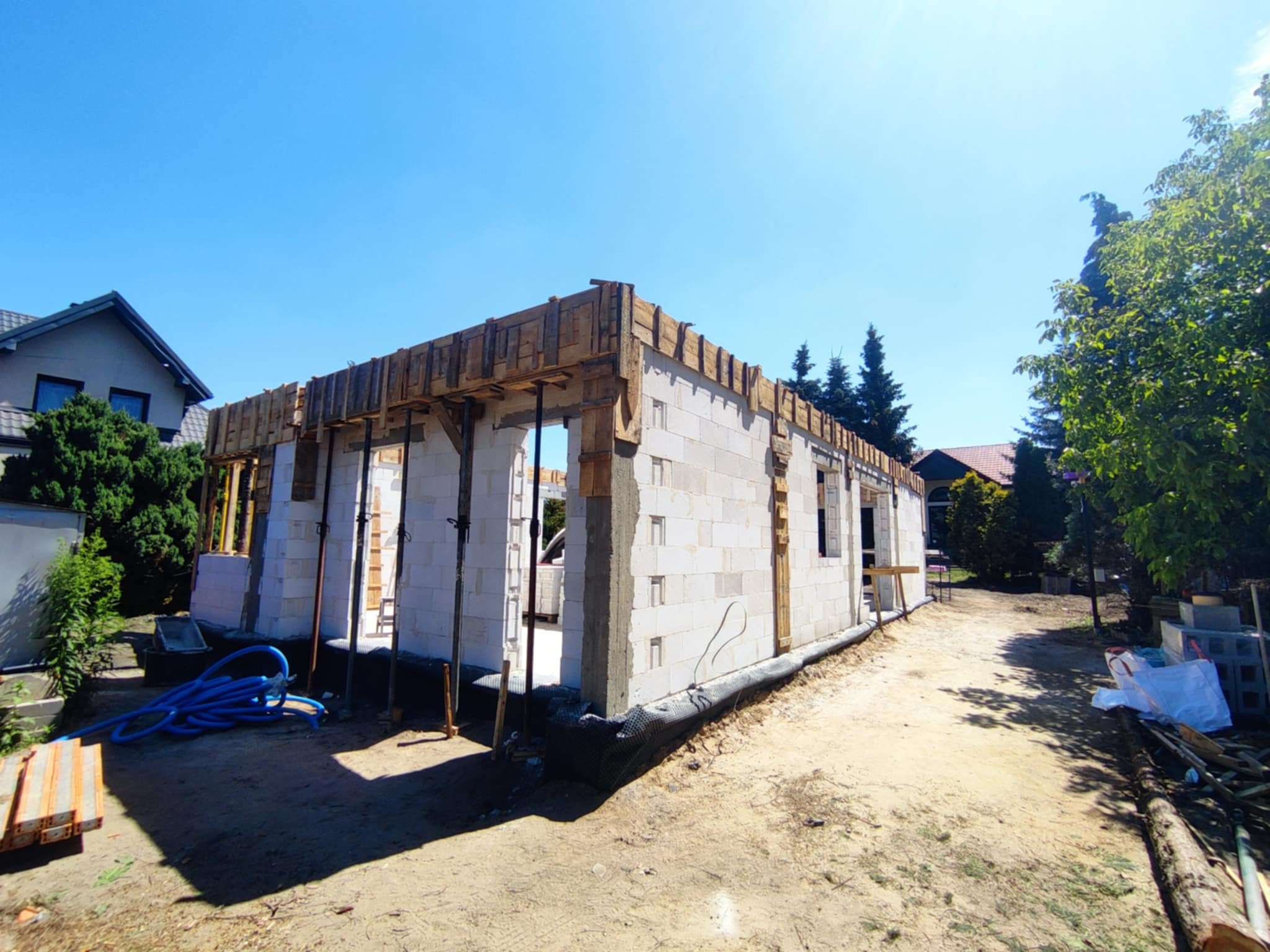 Budowa domu jednorodzinnego: wznoszenie ścian z bloczków betonowych, szalunek drewniany na górze, podpora stalowa. Widok w słoneczny dzień.