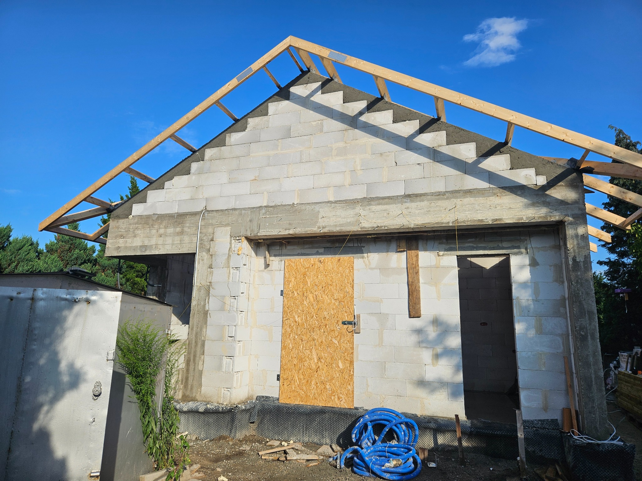 Budowa domu z białych bloczków: surowy stan, widoczne drewniane elementy konstrukcji dachu, niebieskie rury i tymczasowe drzwi z płyty OSB.