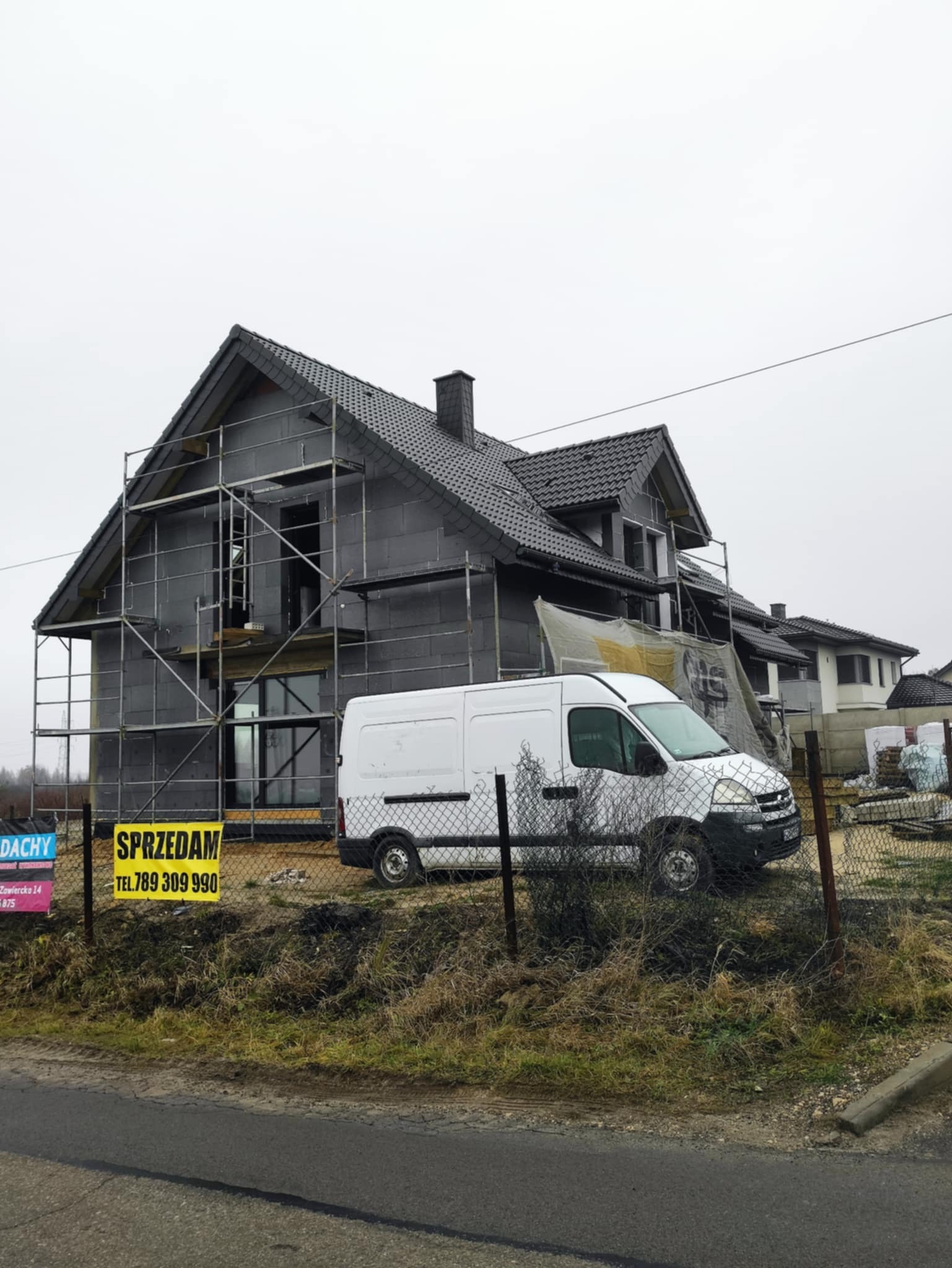 Dom w trakcie ocieplania, obłożony styropianem, z rusztowaniami. Na pierwszym planie biały samochód dostawczy i tablica 'Sprzedam'. Pochmurny dzień.