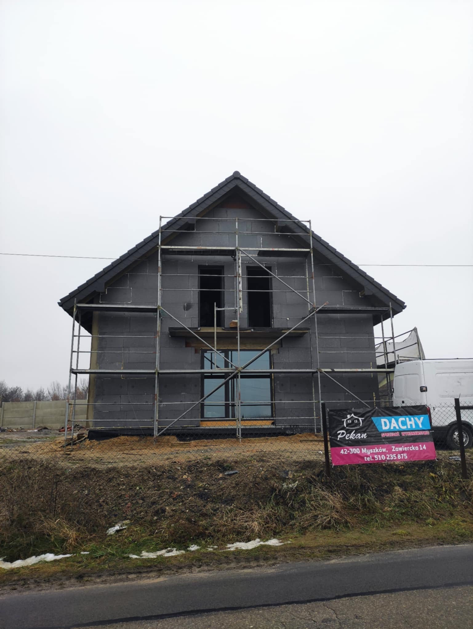 Dom w trakcie ocieplania, pokryty szarym styropianem, otoczony rusztowaniami. Widoczny baner reklamowy firmy dekarskiej. Pochmurne niebo w tle.