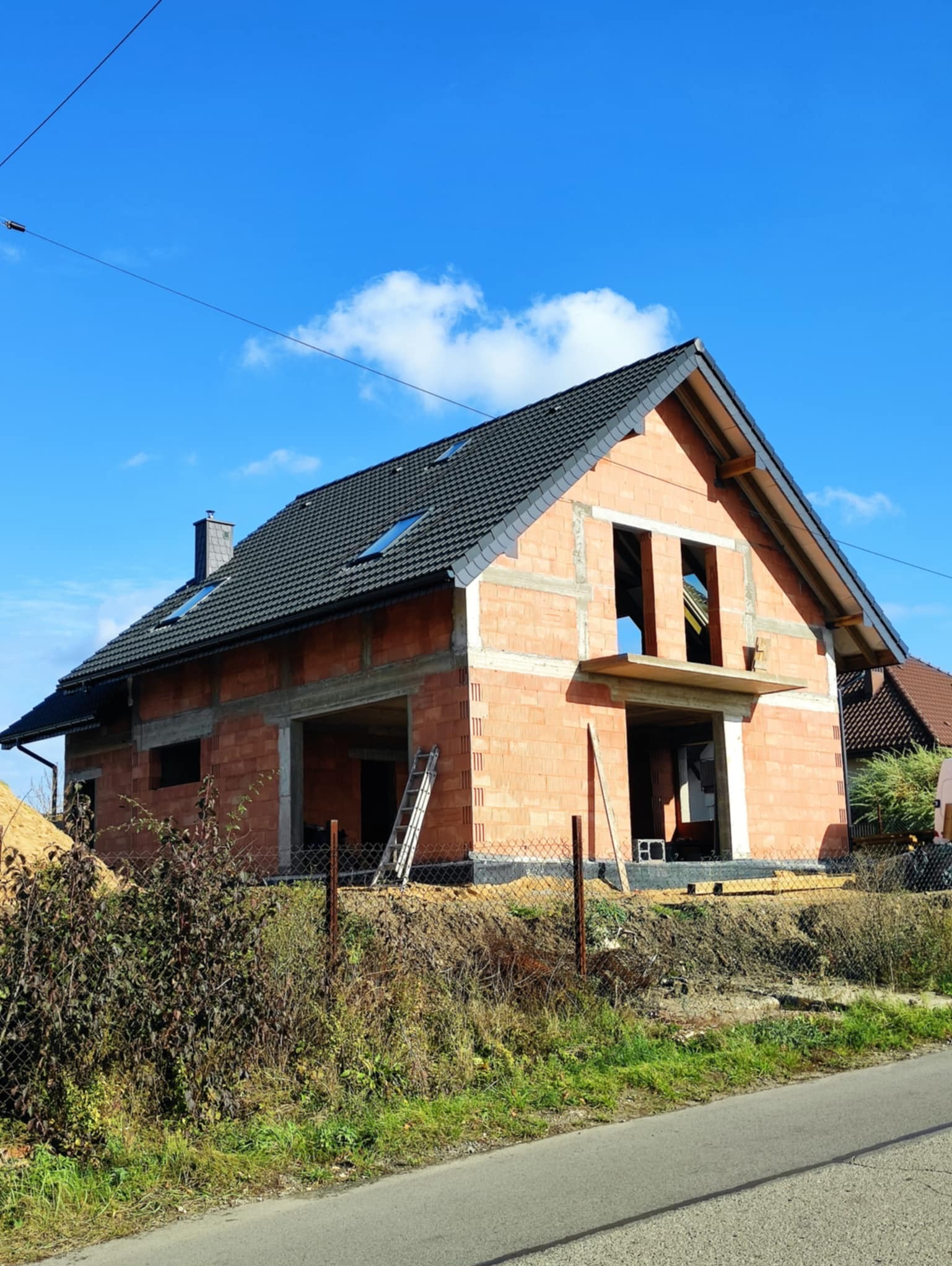 Surowy dom z czerwonej cegły pod błękitnym niebem, z widocznym rusztowaniem i czarnym dachem. Budowa w toku, widok z zewnątrz.