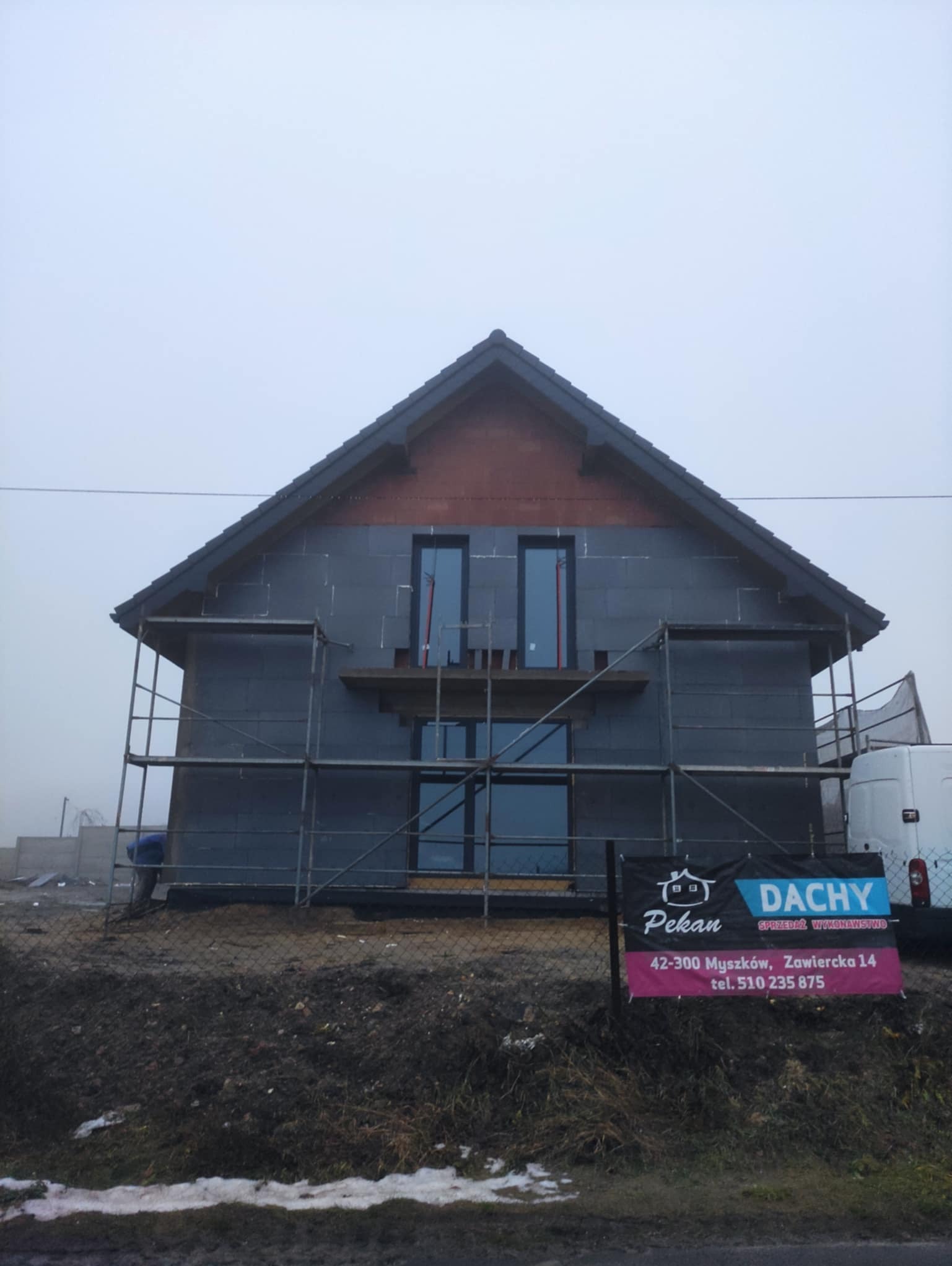 Surowy dom jednorodzinny w trakcie budowy, z widocznym ociepleniem ścian zewnętrznych i rusztowaniami. Na pierwszym planie baner reklamowy firmy.