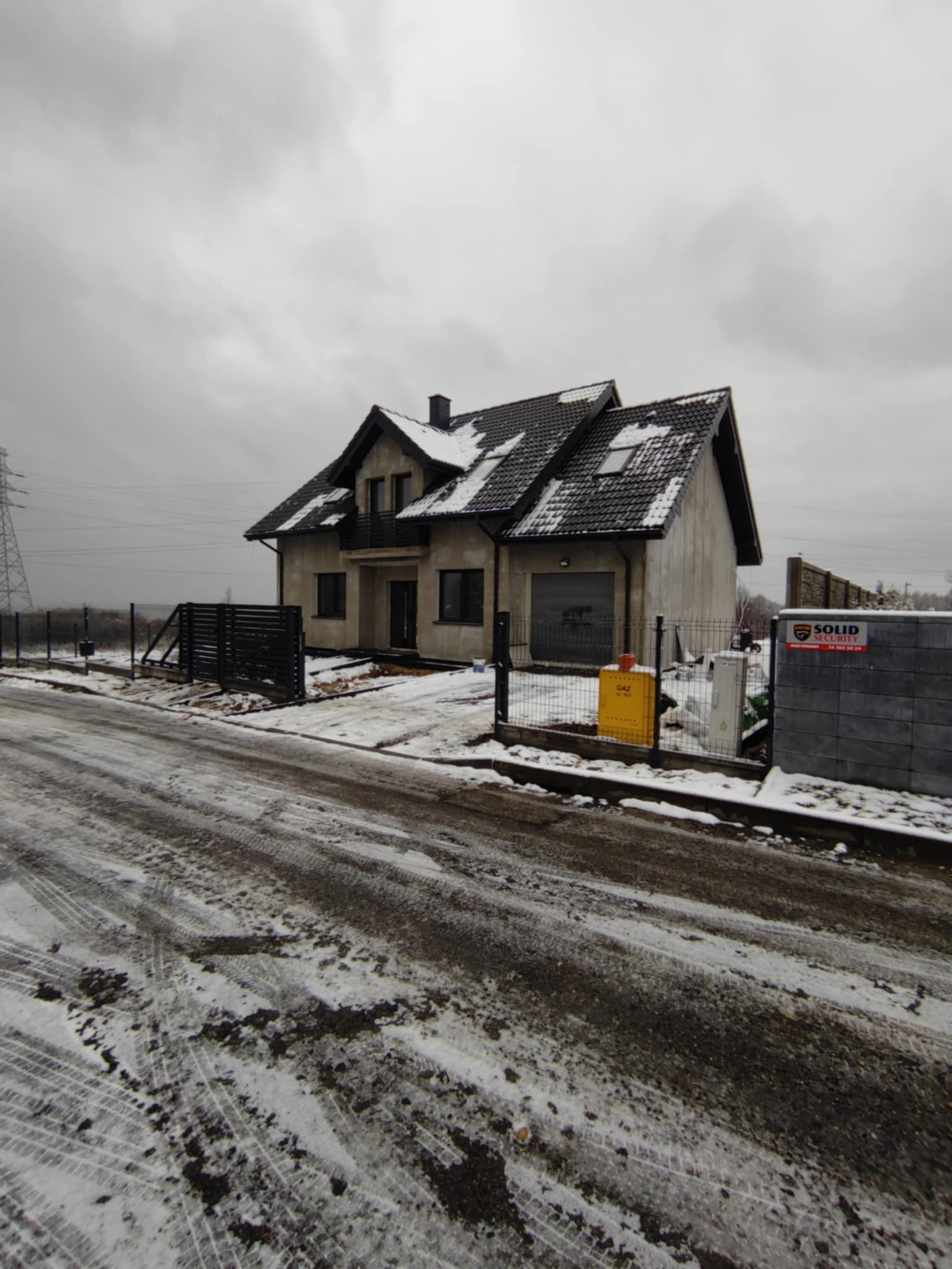 Surowy dom jednorodzinny z ciemną dachówką i szarą elewacją w zimowej scenerii. Widoczne ogrodzenie i skrzynka gazowa. Pochmurne niebo w tle.