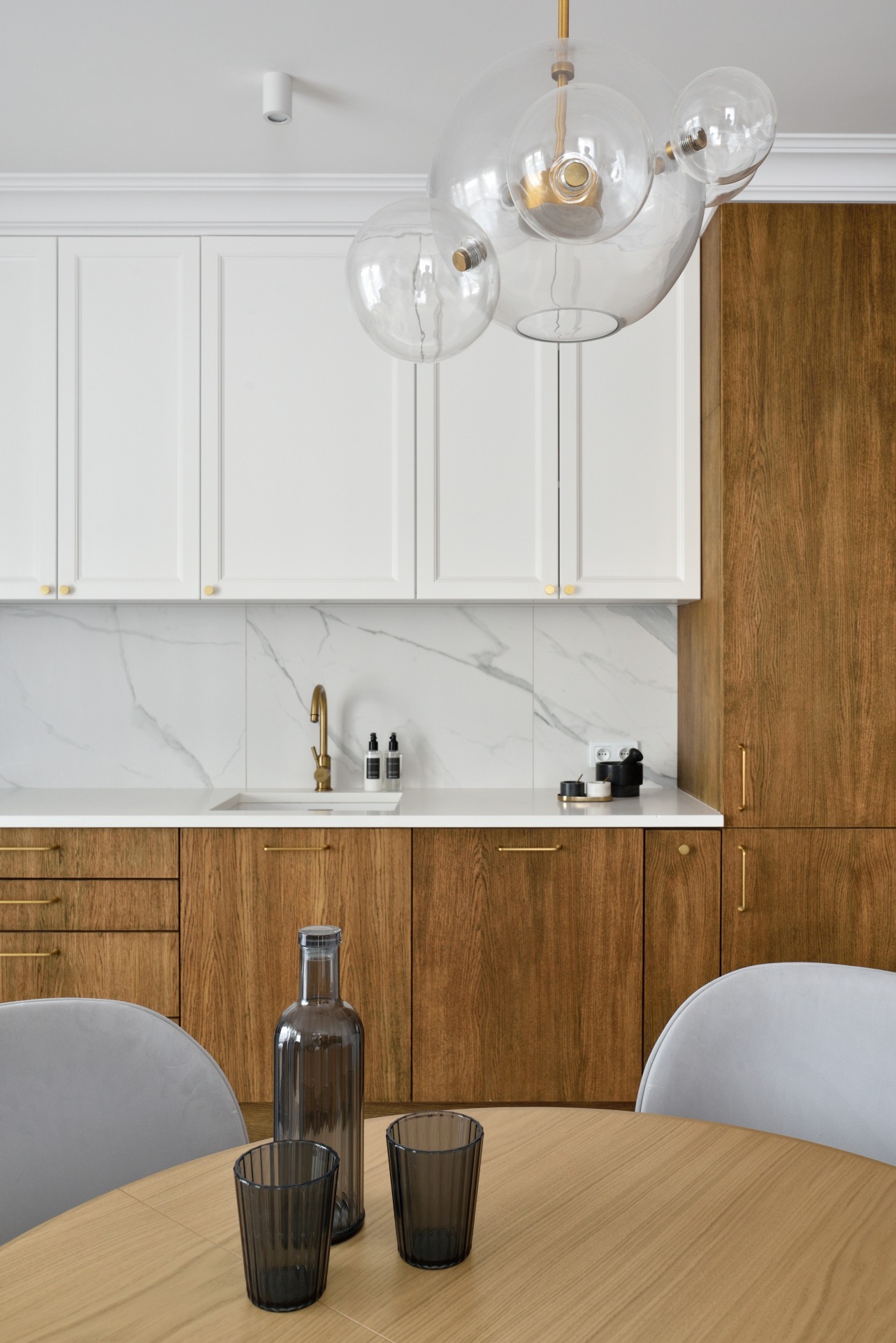 Nowoczesna kuchnia z drewnianymi i białymi szafkami, marmurowym backsplashiem i okrągłym stołem z szarymi krzesłami. Szklana butelka i szklanki na blacie.