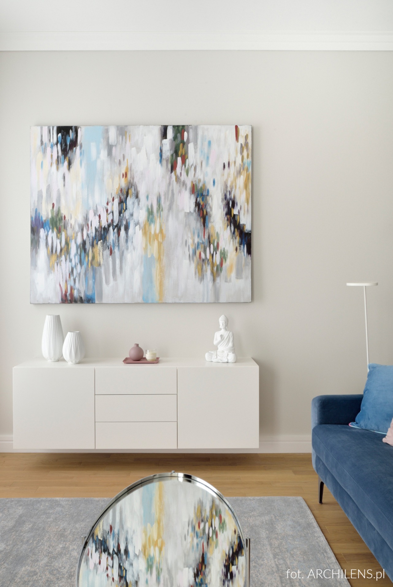 Wnętrze salonu z abstrakcyjnym obrazem nad białą komodą, na której stoją białe wazony i figurka Buddy. Na pierwszym planie stolik kawowy z lustrzanym blatem.