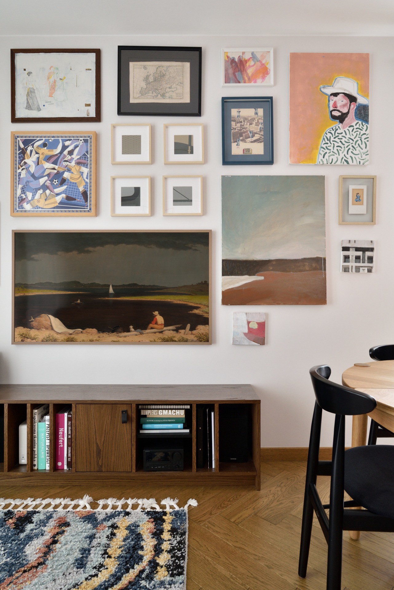 Salon z galerią sztuki na ścianie: różnorodne obrazy w ramach nad drewnianą komodą i wzorzystym dywanem. Krzesło w tle. Unikalna aranżacja wnętrza.