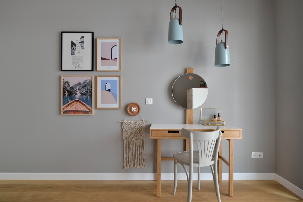 Nowoczesna aranżacja wnętrza z toaletką, okrągłym lustrem, wiszącymi lampami w kolorze pastelowego błękitu oraz galerią plakatów na szarej ścianie.