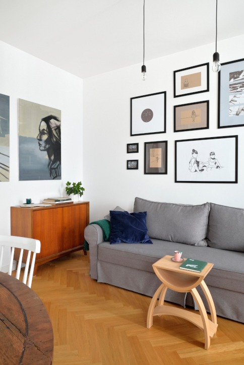 Jasne wnętrze pokoju z szarą sofą, drewnianą komodą z obrazem kobiety, galerią czarno-białych grafik na ścianie i parkietem w jodełkę.