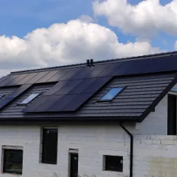 Dom w trakcie budowy z panelami słonecznymi zintegrowanymi z dachem, widoczne okna dachowe i białe ściany z betonu komórkowego.