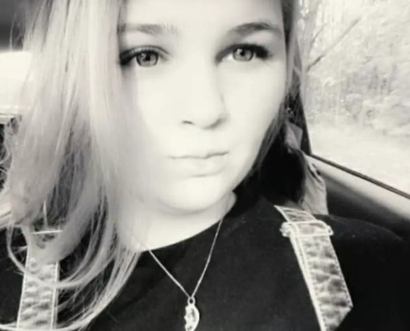 Portret kobiety w czarno-białej tonacji, z delikatnym uśmiechem, srebrnym naszyjnikiem z zawieszką w kształcie połowy serca i włosami opadającymi na ramiona, siedzącej w samochodzie z widocznym...