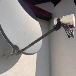 Szara antena satelitarna zamontowana na jasnej ścianie budynku, widoczne kable i elementy mocujące, zadaszenie w tle.