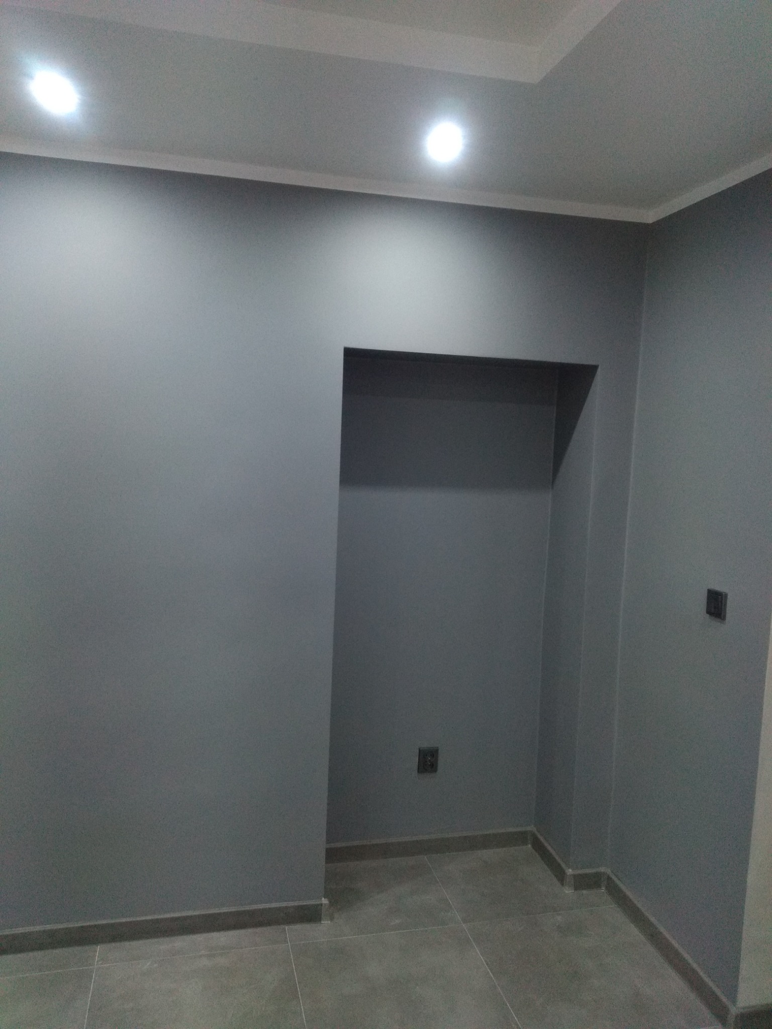 Szare wnętrze pokoju po remoncie, widoczne gładkie ściany pomalowane na szaro, wnęka w ścianie oraz szara podłoga z płytek. Na ścianie widoczny kontakt elektryczny i włącznik światła.