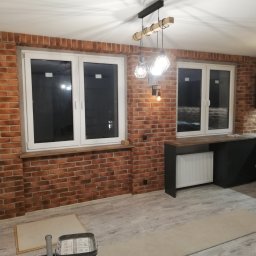 JACEK TIMMLER - Wykończona kuchnia z ceglaną ścianą, białymi oknami i czarnymi szafkami, oświetlona wiszącymi lampami z drewnianymi elementami.