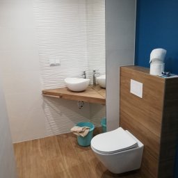 JACEK TIMMLER - Nowoczesna łazienka z umywalką na drewnianym blacie, toaletą podwieszaną obok drewnianej zabudowy, ściany wyłożone białymi płytkami o falistej strukturze i fragmentem ściany pomalowanej na niebiesko.