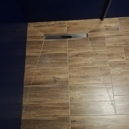 JACEK TIMMLER - Ułożone płytki podłogowe imitujące drewno w łazience, widoczny odpływ liniowy i fragment niebieskiej ściany oraz stopy w butach.