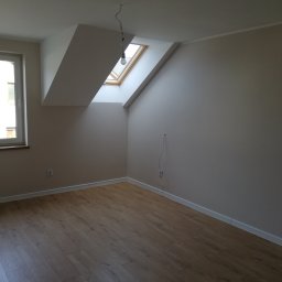 JACEK TIMMLER - Puste, jasne poddasze z oknem dachowym, panele podłogowe w odcieniach brązu, białe ściany przygotowane do malowania, widoczne gniazdka elektryczne i wystające kable.