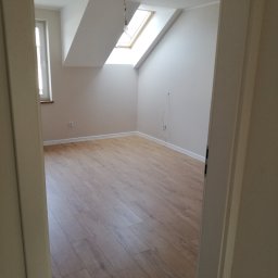 JACEK TIMMLER - Poddasze z oknem dachowym po remoncie, świeżo ułożone panele podłogowe w odcieniach brązu, białe ściany i listwy przypodłogowe, widok zza futryny drzwi.