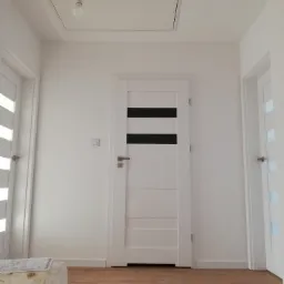 Nowoczesny korytarz po remoncie: białe ściany, panele podłogowe, dwie białe drzwi z czarnymi i przezroczystymi wstawkami, sufit z klapą rewizyjną i wiszącą żarówką.