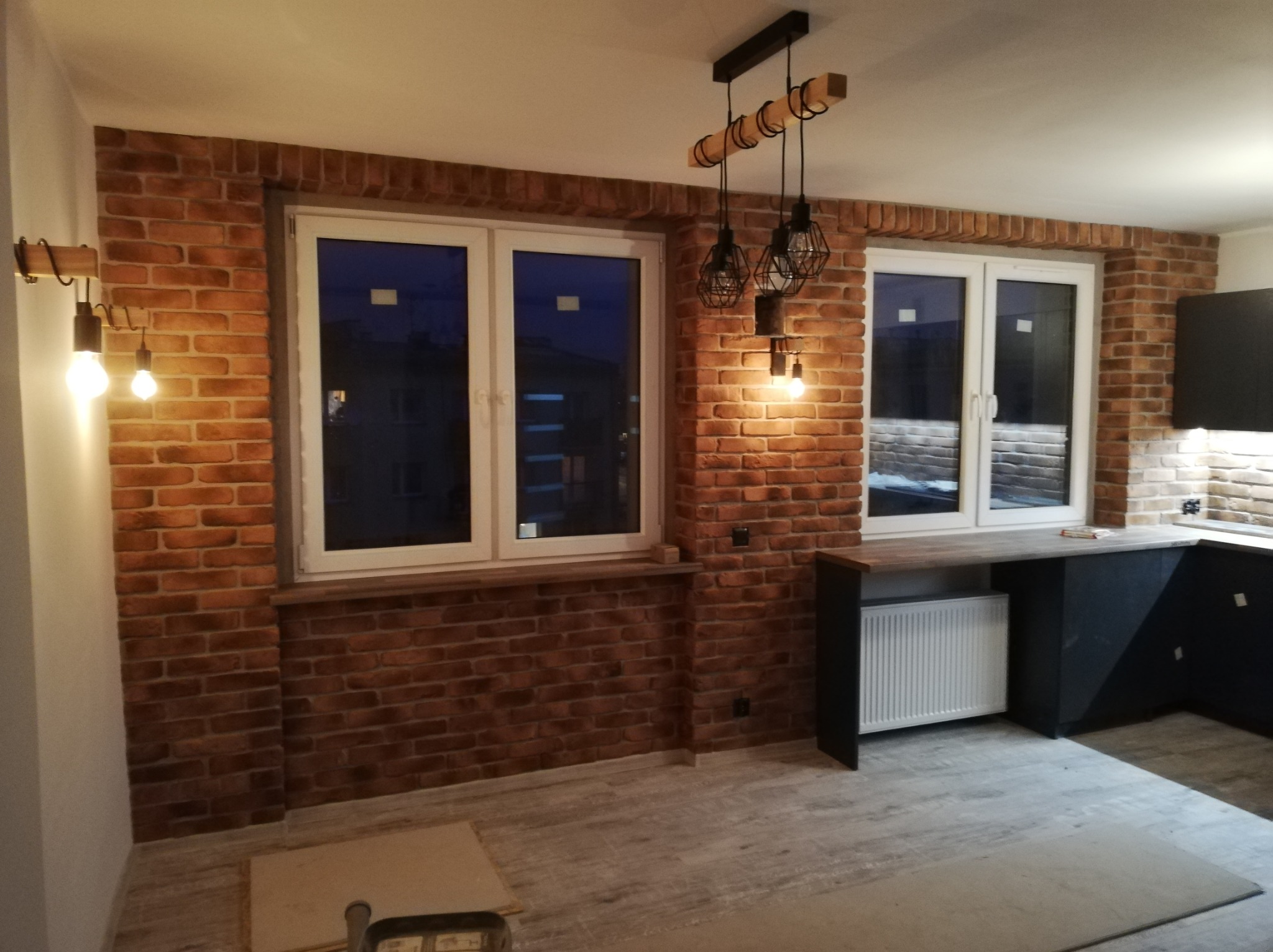 Kuchnia z ceglaną ścianą, oświetleniem w stylu industrialnym i szarymi panelami podłogowymi, widok na dwa okna z białymi ramami.