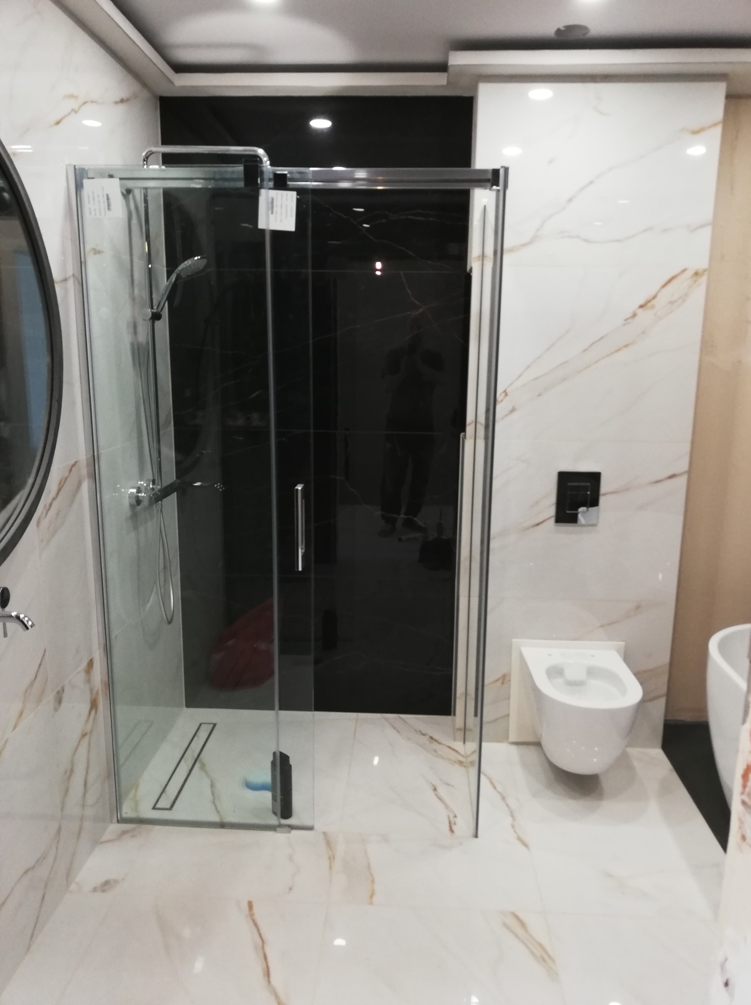 Nowoczesna kabina prysznicowa z czarnymi drzwiami i marmurowymi płytkami, widoczny odpływ liniowy, wisząca toaleta i fragment wanny.