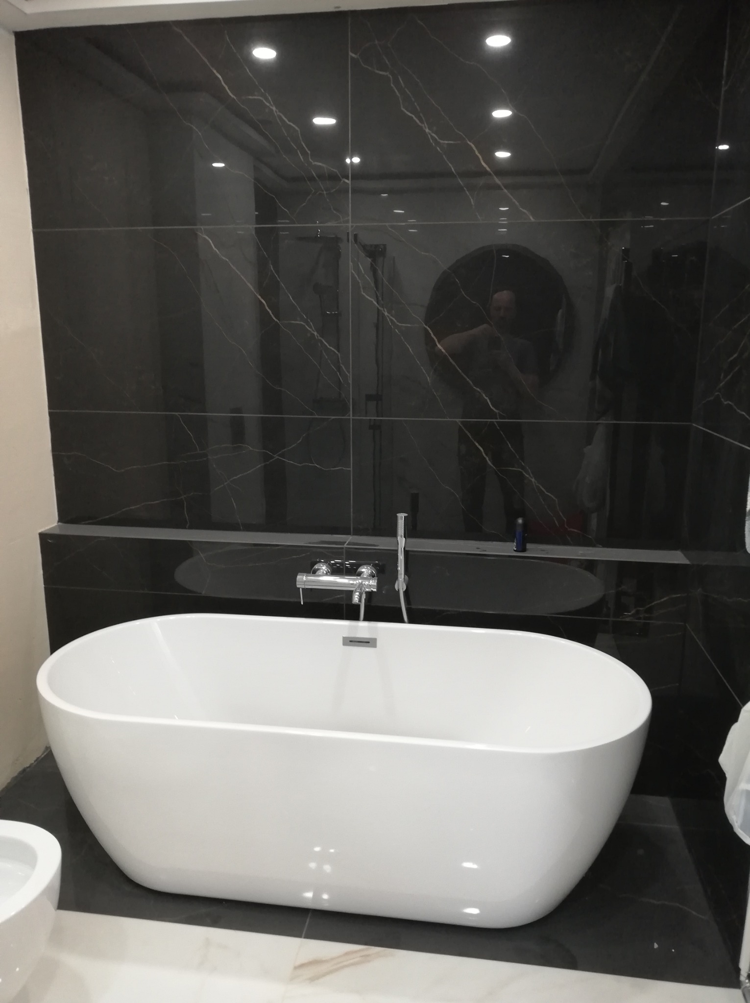 Nowoczesna łazienka z wolnostojącą białą wanną na tle czarnych płytek imitujących marmur z widocznym odbiciem osoby w lustrze.