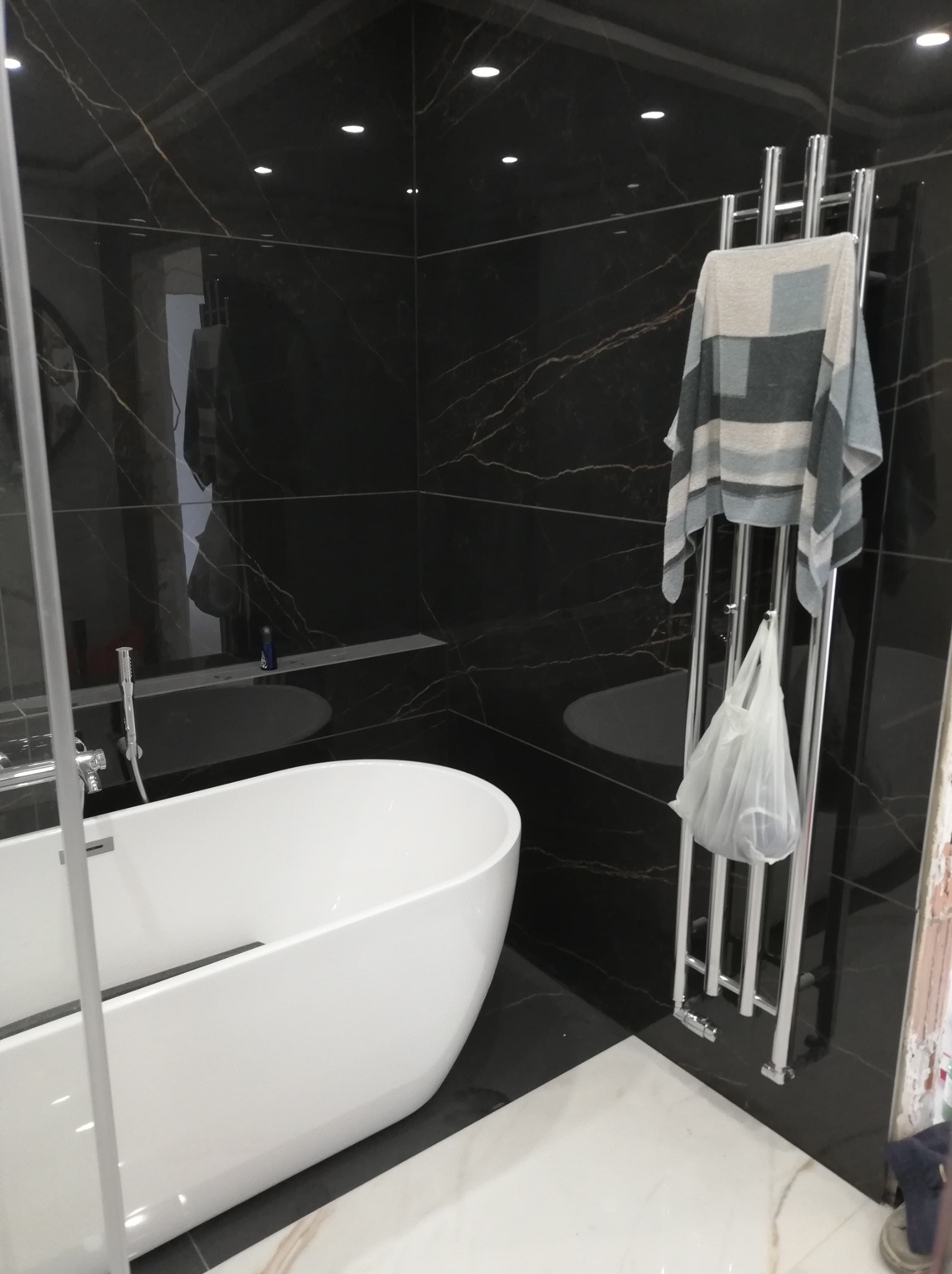 Nowoczesna łazienka z wolnostojącą białą wanną, czarnymi płytkami imitującymi marmur z wyraźnym żyłkowaniem i grzejnikiem drabinkowym z powieszonym ręcznikiem w odcieniach szarości.