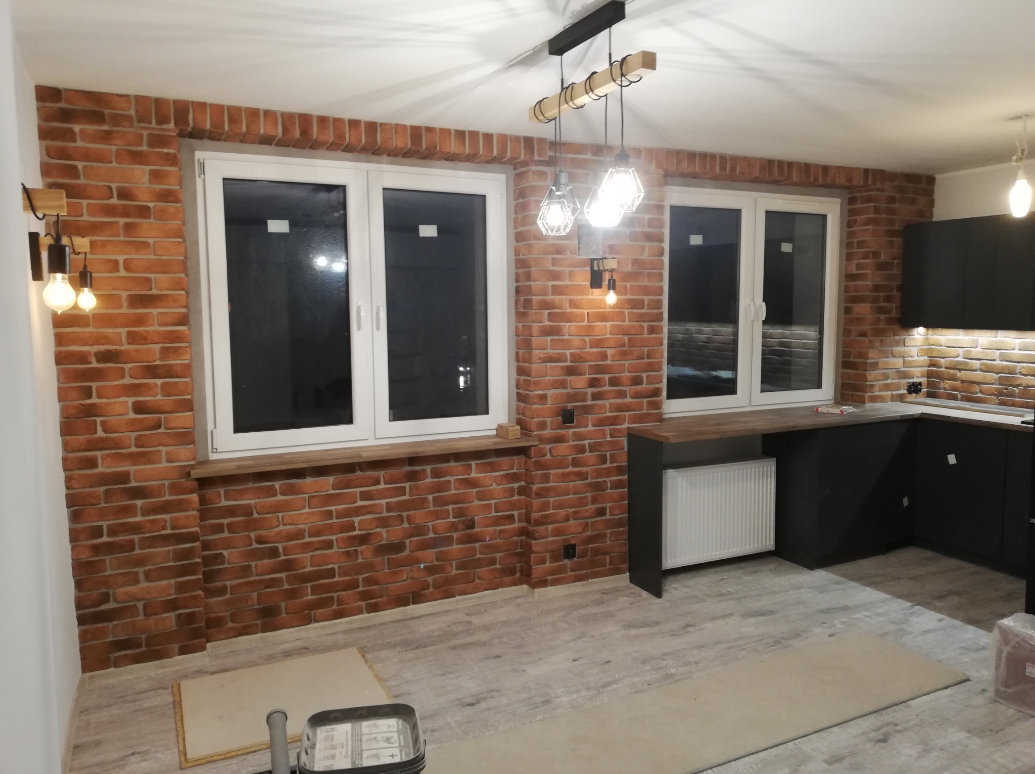 Wykończona kuchnia z ceglaną ścianą, białymi oknami i czarnymi szafkami, oświetlona wiszącymi lampami z drewnianymi elementami.