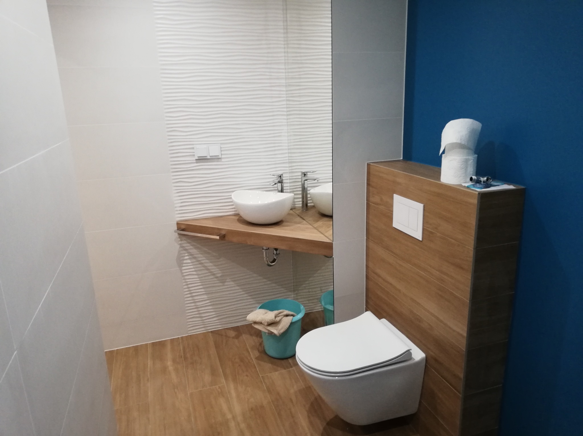Nowoczesna łazienka z umywalką na drewnianym blacie, toaletą podwieszaną obok drewnianej zabudowy, ściany wyłożone białymi płytkami o falistej strukturze i fragmentem ściany pomalowanej na niebiesko.