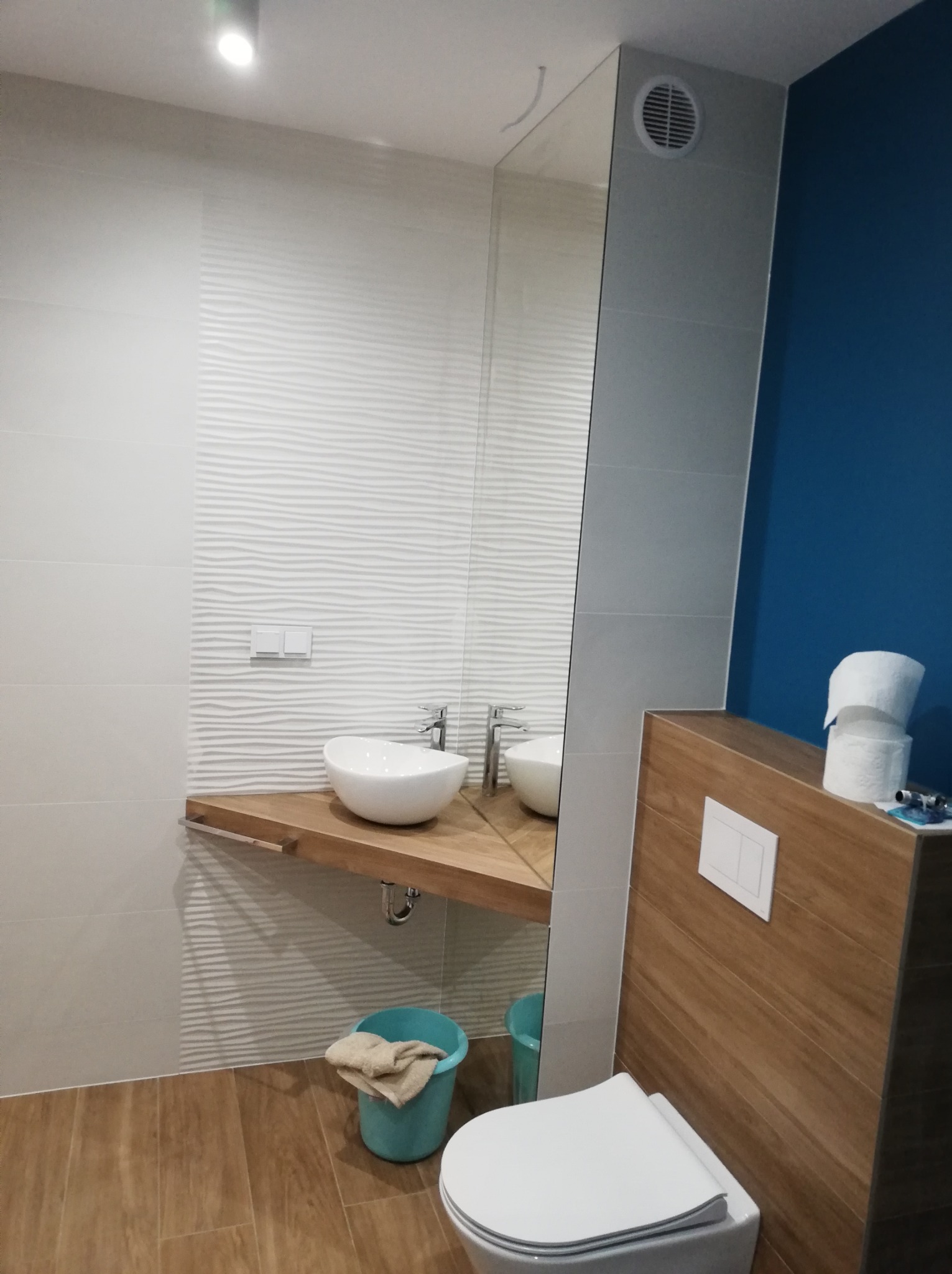 Nowoczesna łazienka z umywalką nablatową na drewnianym blacie, toaleta z zabudową w odcieniach drewna, błękitna ściana w tle i podłoga imitująca drewno.