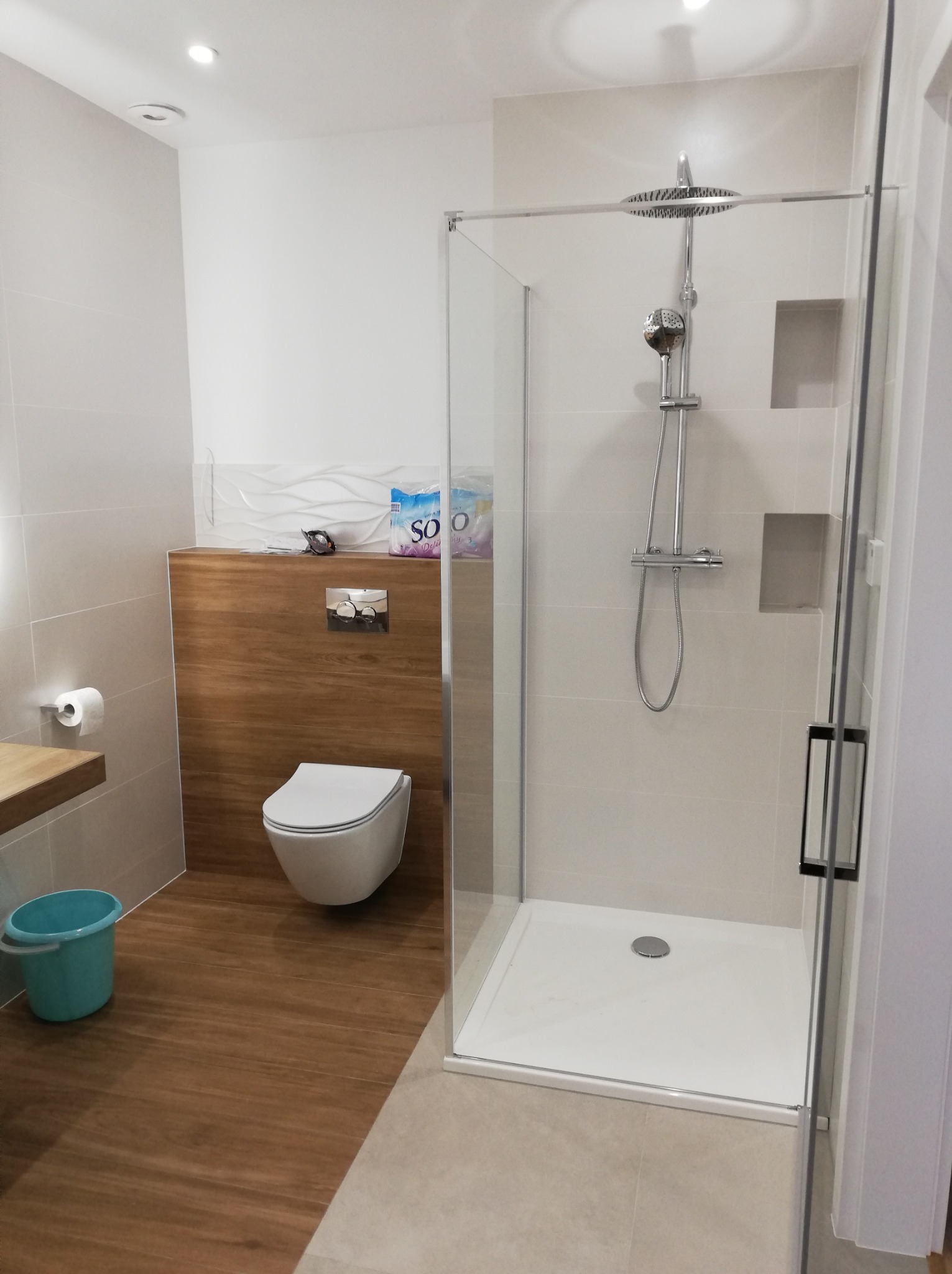 Nowoczesna łazienka z kabiną prysznicową typu walk-in, podwieszaną toaletą, drewnopodobną zabudową i turkusowym wiaderkiem.